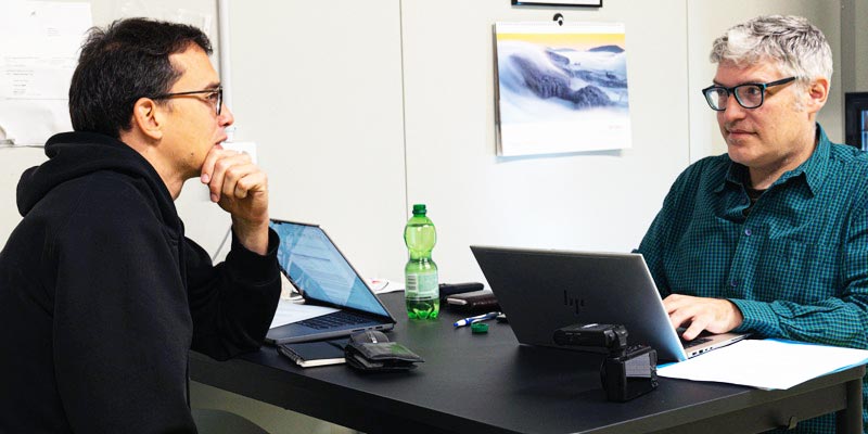 Zwei Männer sitzen an einem Tisch, arbeiten an Laptops und führen ein Gespräch. Im Hintergrund ist ein Kalender mit einem Naturmotiv zu sehen. Auf dem Tisch liegen eine Wasserflasche, Notizen und Arbeitsmaterialien.